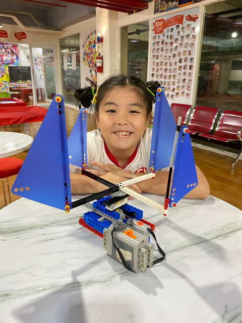 A smiling student resting her chin behind a LEGO model with blue sails and a motor