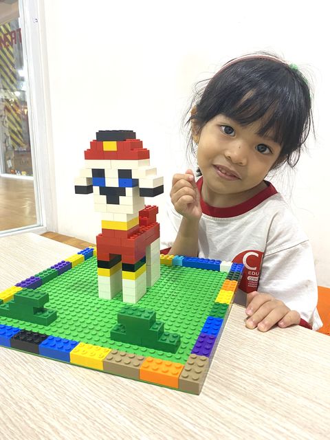 A young girl beside a character she built from LEGO bricks on a green baseplate